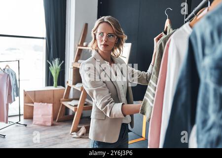 Choisir la meilleure option. Jeune femme attrayante dans des vêtements décontractés élégants choisissant des vêtements du rang et regardant loin tout en se tenant debout dans l'atelier Banque D'Images