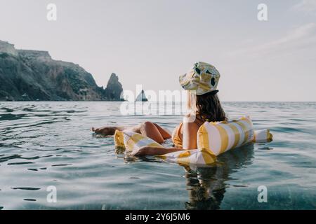 Femme Beach gonflable détente sur un flotteur gonflable rayé dans l'océan près d'une côte rocheuse, portant un chapeau et profitant du soleil. Banque D'Images