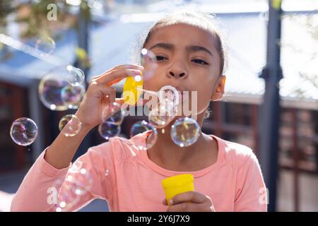 Souffler des bulles, fille afro-américaine profitant de l'activité de plein air dans l'aire de jeux de l'école avec des amis Banque D'Images