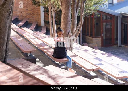 Assise sur le banc, fille afro-américaine en uniforme scolaire regardant à l'intérieur du sac à dos à l'extérieur Banque D'Images