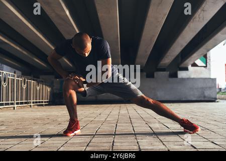 Je m'étire bien. Beau jeune homme africain dans des vêtements de sport s'étirant tout en s'échauffant à l'extérieur Banque D'Images