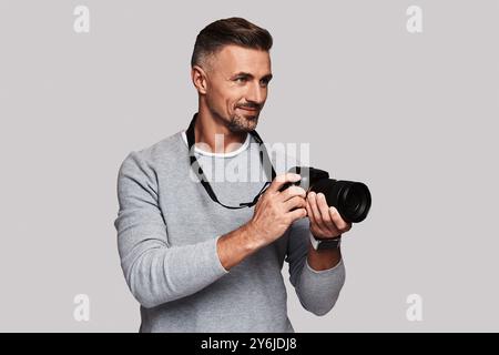 Beau journaliste. Beau jeune homme tenant un appareil photo numérique et souriant tout en se tenant debout sur fond gris Banque D'Images