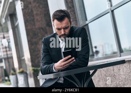 Prendre tout au sérieux. Beau jeune homme en costume complet en utilisant son téléphone intelligent tout en étant assis dans le café à l'extérieur Banque D'Images