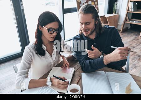 En passant en revue les détails. Vue de dessus de deux jeunes gens modernes en tenue décontractée élégante écrivant quelque chose tout en étant assis dans le bureau créatif Banque D'Images