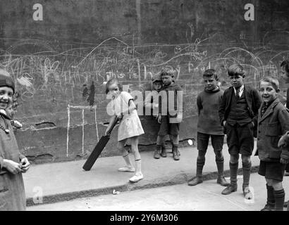 Scènes des bidonvilles de Londres. Waterloo, Londres. Enfants jouant au cricket. Fille tenant la batte. Wicket est dessiné à la craie sur le mur. Garçons en uniforme scolaire regardant. Banque D'Images