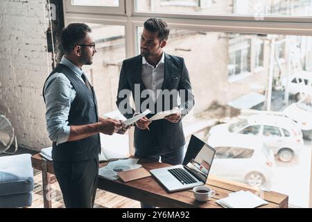 En passant en revue les détails. Vue de dessus de deux jeunes hommes modernes en vêtements de forme travaillant ensemble tout en restant debout à l'intérieur Banque D'Images