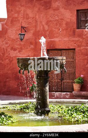 Une belle fontaine en pierre avec de l'eau qui coule dans une cour historique, entourée de murs rouges vibrants et d'une végétation luxuriante. Capture un calme et le temps Banque D'Images