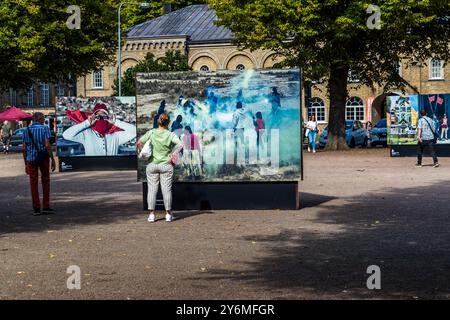 Un visiteur du Landskrona Photo Festival 2024 regarde une image de la série „Shine Heroes“ du photographe uruguayen Federico Estol : des figures se tiennent dans une fumée bleutée sur un terrain découvert, la scène mise en scène comme une action de rue. Un panneau en arrière-plan montre également une figure dont le visage est enveloppé dans un tissu rouge et blanc. Estol a travaillé avec soixante garçons ciriers de La Paz et El Alto en Bolivie, qui ont aidé à mettre en scène les scènes en tant que producteurs et protagonistes d’un essai photographique visant à combattre la stigmatisation sociale attachée à leur métier. Citadellstaden, Skåne, Suède. Banque D'Images