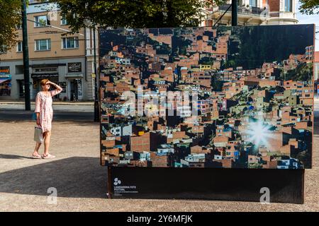 Une passante se protège les yeux en passant devant une grande photographie de la série „Shine Heroes“ de l'artiste uruguayen Federico Estol au Festival de la photo de Landskrona 2024, montrant un quartier de colline densément construit de La Paz ou El Alto, en Bolivie. Estol a travaillé avec soixante garçons chaussures des deux villes, qui ont aidé à mettre en scène les scènes en tant que producteurs et protagonistes d'un essai photographique visant à combattre la stigmatisation sociale attachée à leur métier. Citadellstaden, Skåne, Suède. Banque D'Images