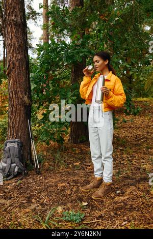 Dans une belle forêt d’automne, une jeune afro-américaine prend un moment pour siroter une boisson chaude pendant sa randonnée. Banque D'Images