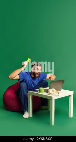 Jeune homme émotionnel et excité regardant sur ordinateur portable, tenant une carte de crédit et exprimant l'excitation sur fond de studio vert Banque D'Images