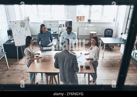 En passant en revue les détails. Vue de dessus de jeunes gens modernes en tenue décontractée intelligente discutant des affaires tout en se tenant derrière le mur de verre dans la salle du conseil Banque D'Images