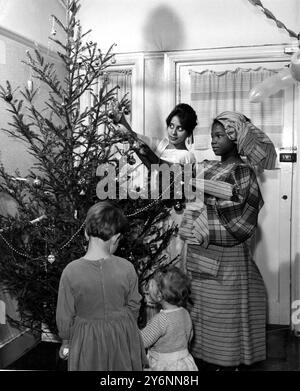 Vêtus de leurs costumes nationaux, Fatima Chakera (à gauche) de Dar es Salaam, Tanganyika, étudiante au Norwood Technical College, et Funlayo Odufuwq, originaire du Nigeria occidental, étudiante au Croydon Technical College, décorent le sapin de Noël dans le hall de la maison de Streatham Road, Londres SW16 de la famille Norgate. Ils sont deux d'un groupe d'étudiants étrangers qui doivent passer Noël avec M. Bernard Norgate, un cadre d'une entreprise d'emballage de viande Smithfield, sa femme Daphne et leurs deux enfants - vu sur la photo - Deborah 4 et Miriam 16 mois. - 18 décembre 1961 Banque D'Images