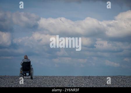 Le Hourdel, France - 09 17 2024 : vue arrière d'un homme en fauteuil roulant sur la plage de galets regardant la mer Banque D'Images