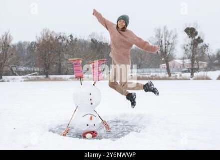 bonhomme de neige drôle se tient à l'envers sur un lac enneigé gelé, une jeune femme de bonne humeur saute près de lui Banque D'Images