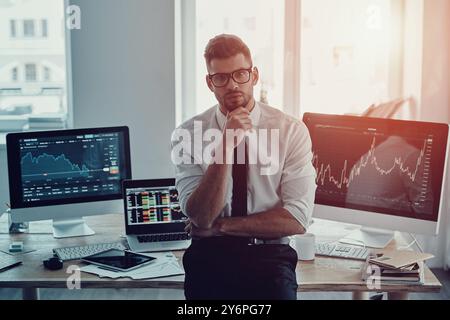 Homme d'affaires moderne. Beau jeune homme en vêtements de forme gardant la main sur le menton et regardant la caméra tout en se tenant debout dans le bureau Banque D'Images