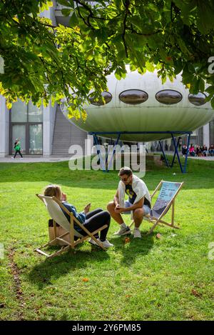 Un couple est assis dans des transats sur la pelouse devant la Pinakothek der moderne et l'installation artistique Futuro House de Matti Suuronen, Munich Banque D'Images