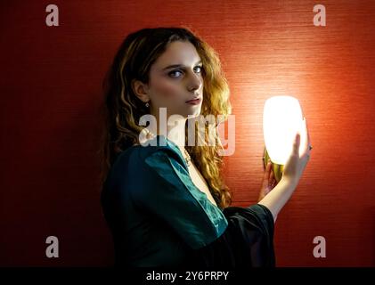 La charmante mannequin Ava Nicholson est assise à côté d'une lumière de chevet lumineuse à l'intérieur d'une chambre à l'hôtel Dundee Queens posant pour des photographies en Écosse Banque D'Images