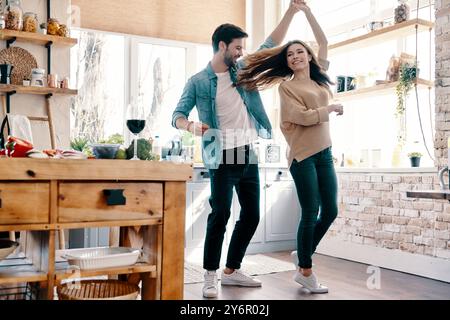 Deux cœurs remplis d'amour. Pleine longueur de beau jeune couple dans des vêtements décontractés dansant et souriant tout en se tenant dans la cuisine à la maison Banque D'Images