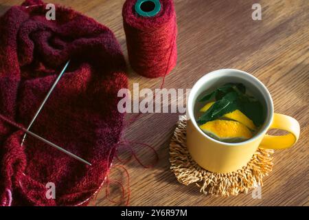 Citron et feuille de menthe dans une tasse jaune et tricot rouge foncé avec des aiguilles à tricoter. Style de vie automnal. Fil de laine couleur cerise avec tisane sur table. Banque D'Images