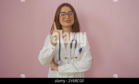 Médecin hispanique confiant avec des lunettes, pointant vers le haut avec une expression sérieuse, porte une blouse de laboratoire blanche et un stéthoscope, posant contre un EI Banque D'Images