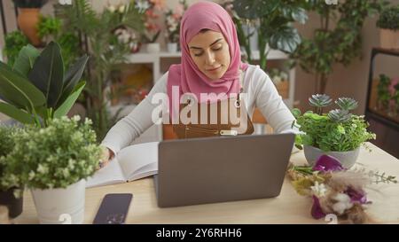 Une jeune femme avec un hijab engagée dans la lecture d'un livre sur son ordinateur portable dans une pièce remplie de plantes, symbolisant l'étude ou le travail à domicile. Banque D'Images