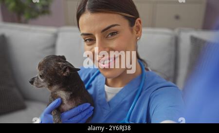 Une jeune femme souriante dans un haut bleu tient un chihuahua dans un salon confortable, prenant un selfie Banque D'Images