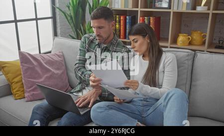 Couple hispanique examinant des documents ensemble sur un canapé avec un ordinateur portable dans un intérieur confortable de salon. Banque D'Images