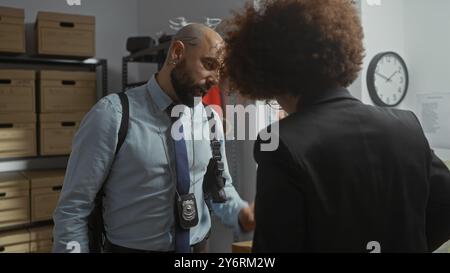 Un homme et une femme en tenue professionnelle conversent dans un poste de police rempli de boîtes à preuves. Banque D'Images