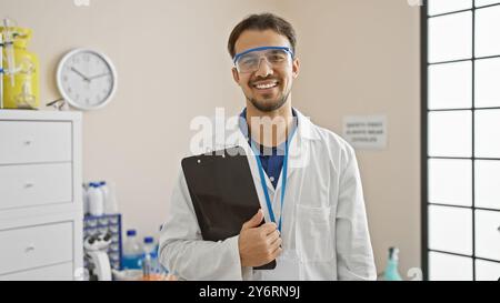 Jeune homme hispanique avec la barbe portant une blouse de laboratoire et des lunettes de sécurité, tenant une presse-papiers dans le laboratoire Banque D'Images