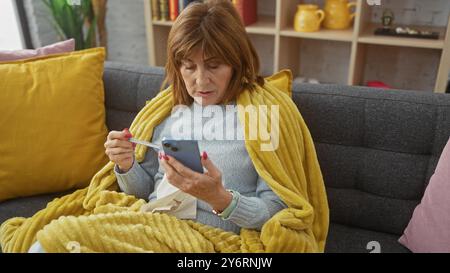 Une femme d'âge moyen à la maison utilise un smartphone enveloppé dans une couverture jaune confortable sur le canapé. Banque D'Images