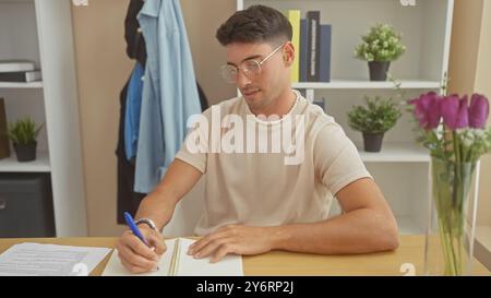 Bel homme hispanique étudiant des documents à une table à la maison avec des fleurs dans un vase et des étagères en arrière-plan. Banque D'Images