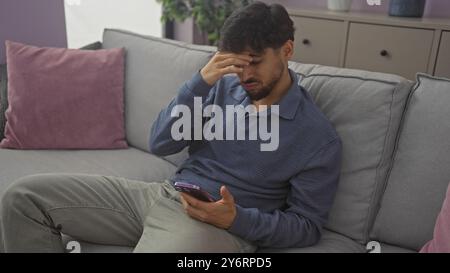 Un beau jeune homme avec une barbe semble stressé tout en tenant son smartphone sur un canapé à l'intérieur. Banque D'Images
