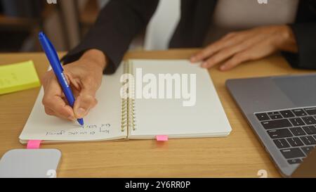 Femme d'âge moyen écrivant dans un cahier au bureau à domicile avec ordinateur portable et notes adhésives Banque D'Images