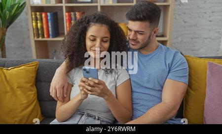 Un jeune couple est assis près d'un canapé à la maison, regardant heureux un smartphone ensemble dans un salon confortable. Banque D'Images