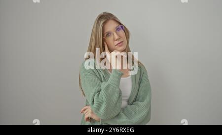 Une jeune femme réfléchie avec des lunettes et un pull pose sur un fond blanc isolé, dégageant une beauté décontractée. Banque D'Images