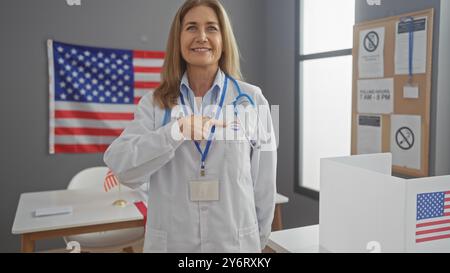 Un professionnel de la santé d'une femme d'âge moyen confiante pointe sur son autocollant « j'ai voté » à l'intérieur avec un drapeau américain en toile de fond. Banque D'Images