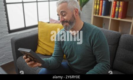 Homme barbu mature sourit tout en tenant une tablette dans un cadre intérieur confortable avec étagère et décor moderne. Banque D'Images