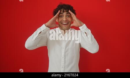 Un homme hispanique surpris dans une chemise blanche pose avec une expression excitée sur un fond rouge vif. Banque D'Images