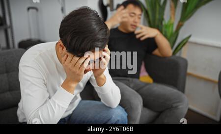 Deux hommes asiatiques discutent émotionnellement dans un salon moderne, l'un faisant des gestes tandis que l'autre est stressé. Banque D'Images