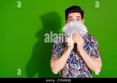 Photo de gars timide étonné avec chemise imprimée habillée de coupe de cheveux élégante cachant le visage derrière des dollars isolé sur fond de couleur verte Banque D'Images