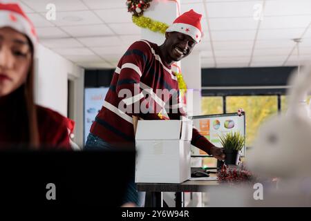 Joyeux employé afro-américain portant chapeau de Noël déballant la boîte de biens, décorant le nouveau bureau après avoir été embauché. Homme d'affaires commençant le travail, excité de travailler dans le bureau orné festif Banque D'Images