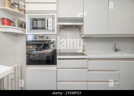 Cuisine meublée avec des meubles blancs, comptoirs, réfrigérateur double, colonne avec fours, plaque vitrocéramique et carrelage blanc. Banque D'Images