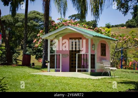 Une petite maison rose avec un toit blanc se trouve dans un champ herbeux. La maison est entourée d'arbres et a un banc en face de lui Banque D'Images