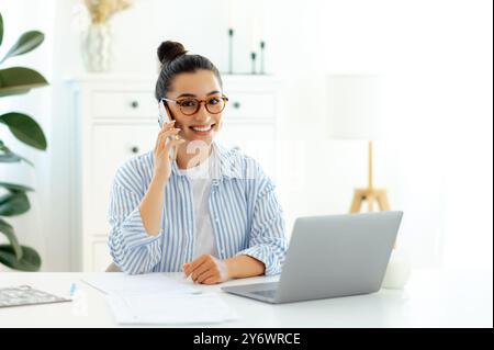 Femme d'affaires caucasienne attrayante réussie parlant par smartphone de stratégie avec un client ou un partenaire tout en travaillant la journée sur son bureau avec un ordinateur portable sur le bureau, sourires amicaux à la caméra Banque D'Images
