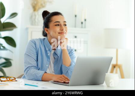 Jeune femme caucasienne heureuse rêveuse dans une chemise bleue, travailleuse de l'entreprise, assise à son lieu de travail avec un ordinateur portable, regardant soigneusement loin et souriant, pensant au profit du projet, rêvant du week-end Banque D'Images