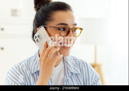 Photo en gros plan de belle femme d'affaires caucasienne réussie avec des lunettes dans une chemise bleue, parlant par smartphone avec un client ou un partenaire tout en travaillant la journée sur son bureau, regardant loin, souriant Banque D'Images