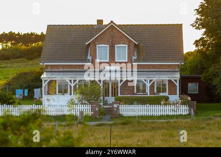 Une maison pittoresque en briques rouges avec une clôture blanche de piquet se trouve paisiblement au milieu des champs verts, luisant dans la lumière chaude du coucher du soleil, créant un co tranquille Banque D'Images