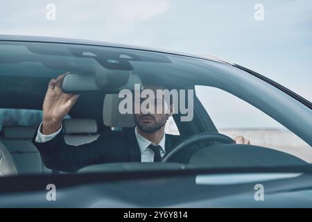 Vérifier chaque détail. Beau jeune homme en costume complet ajustant le rétroviseur tout en conduisant une voiture Banque D'Images