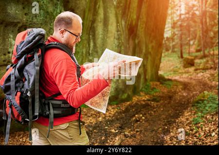 Homme touristique barbu étudie la carte alors qu'il se tient devant un grand rocher dans la forêt dense. Voyageur avec sac à dos gris et lunettes, planification d'aventure en plein air, randonnée ou route d'escalade. Banque D'Images